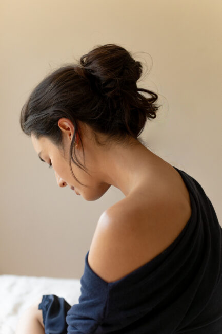 A woman with dark hair in a loose bun sits with her head tilted down, wearing an off-the-shoulder dark top against a neutral background.