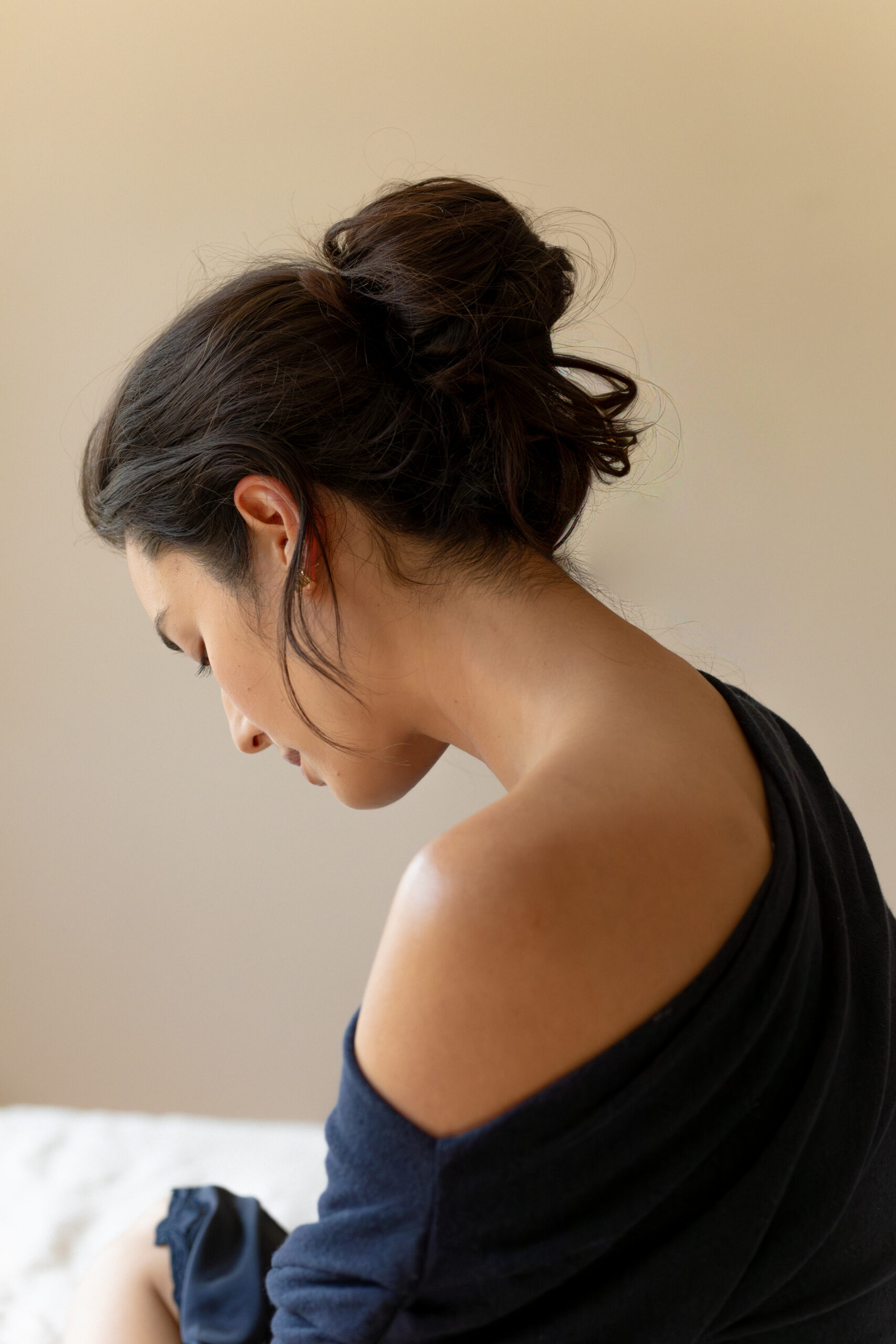 A woman with dark hair in a loose bun sits with her head tilted down, wearing an off-the-shoulder dark top against a neutral background.