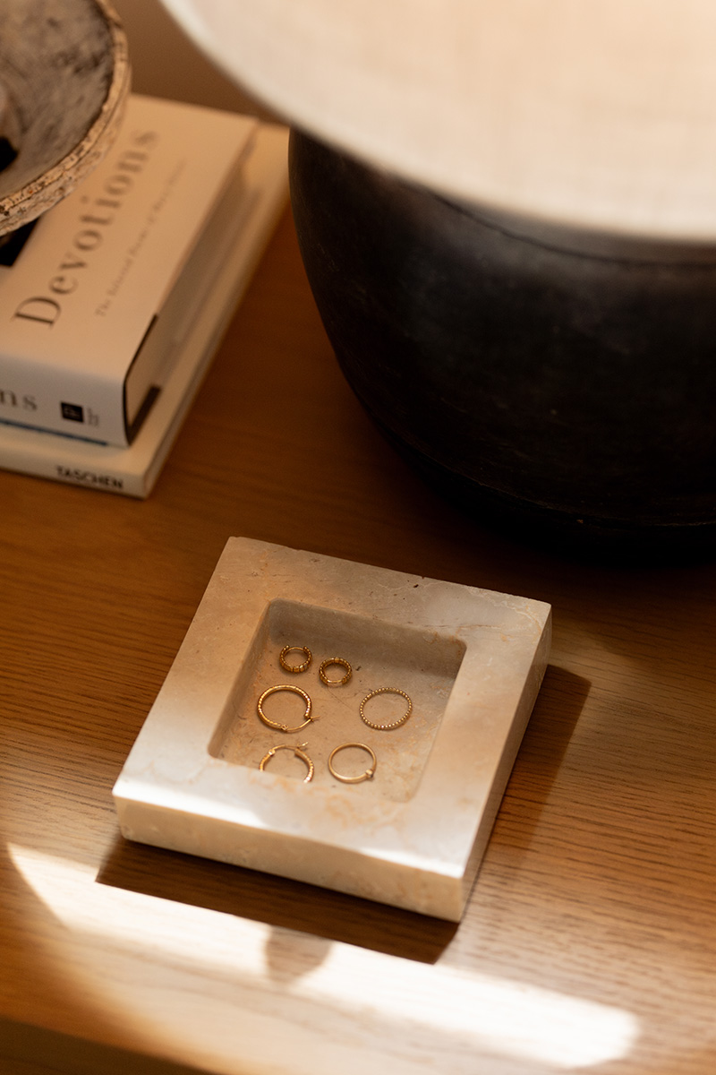 A marble tray holding several gold rings and earrings sits on a wooden surface next to a black pot and a stack of books, including one titled "Devotions.