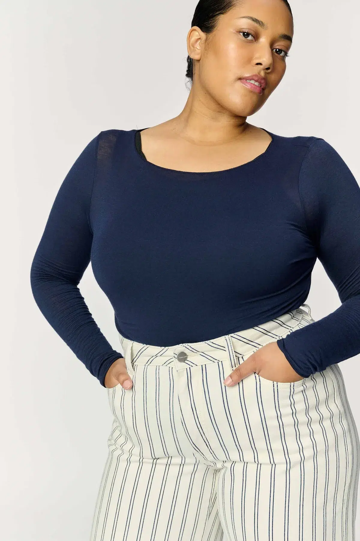 A person stands against a plain background wearing a long-sleeved navy top and high-waisted white pants with vertical black stripes. Hands are in the pockets, and they are looking slightly to the side.