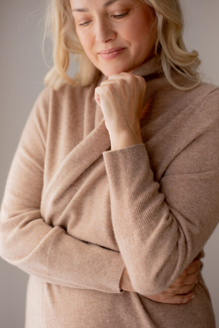 Woman in a beige turtleneck sweater stands with one arm crossed and the other hand resting under her chin, gazing slightly downward with a calm expression.