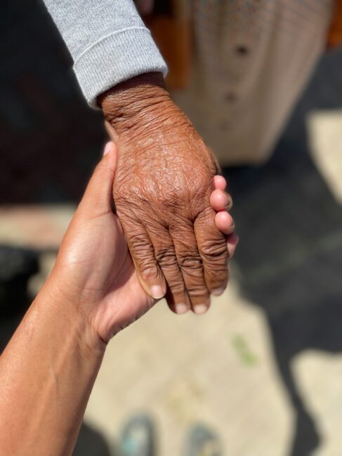 A younger hand gently holds an older hand outdoors, showing a contrast in skin texture and age.