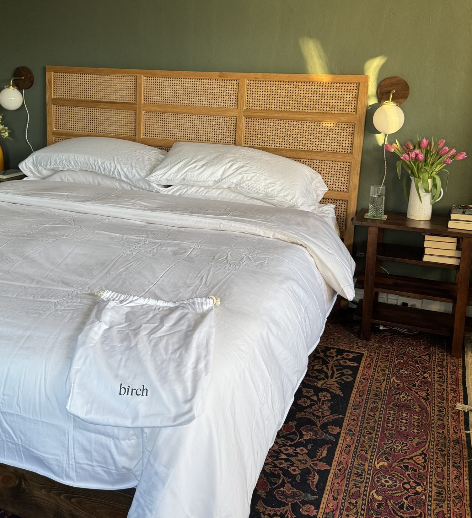 Bedroom with a double bed, white linens, and a rattan headboard. There are two bedside tables, flowers, a yellow vase, and a patterned rug.
