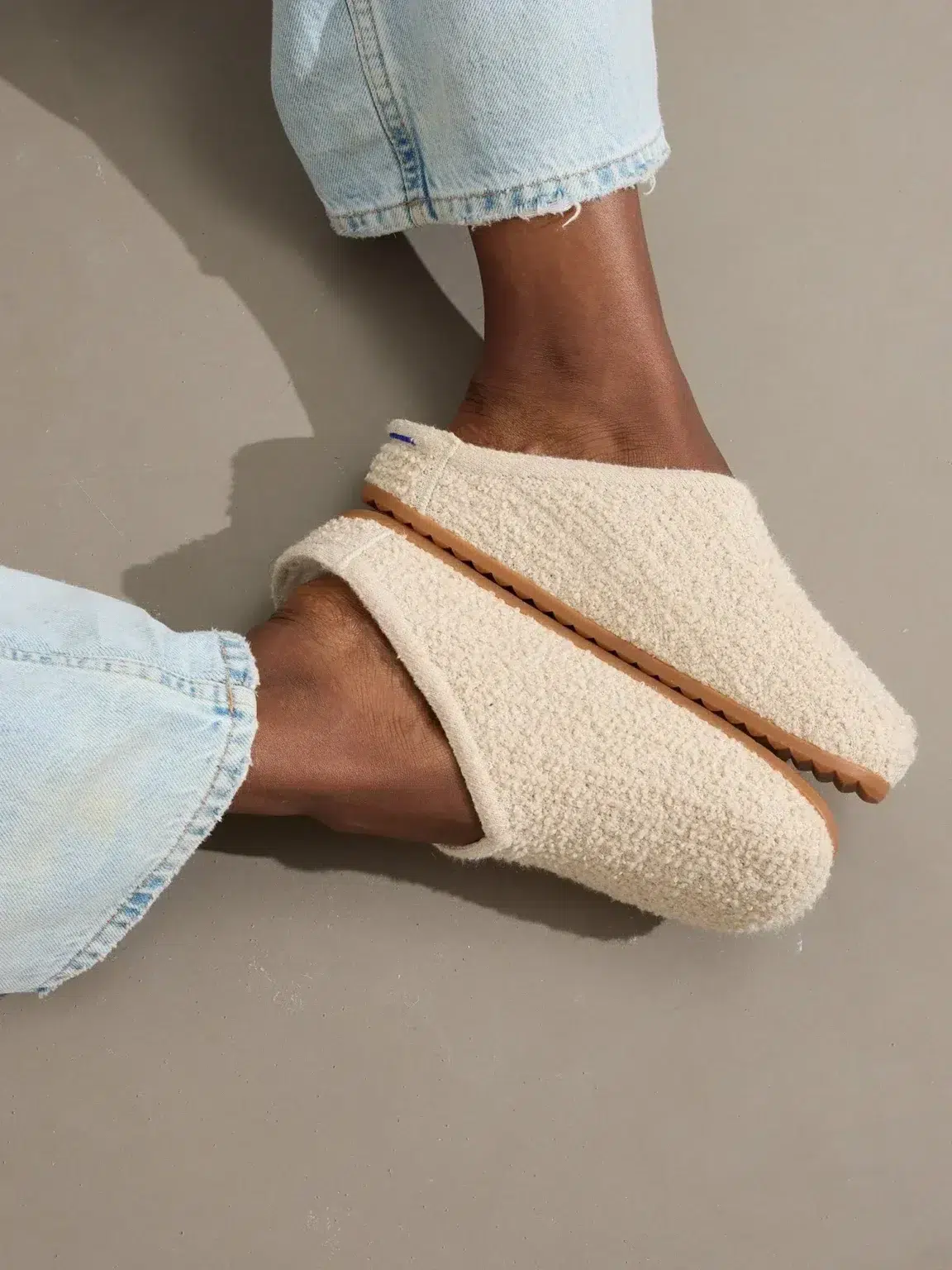 Person wearing light blue jeans and cozy, cream-colored slippers with textured fabric and rubber soles, resting feet on a neutral gray floor.