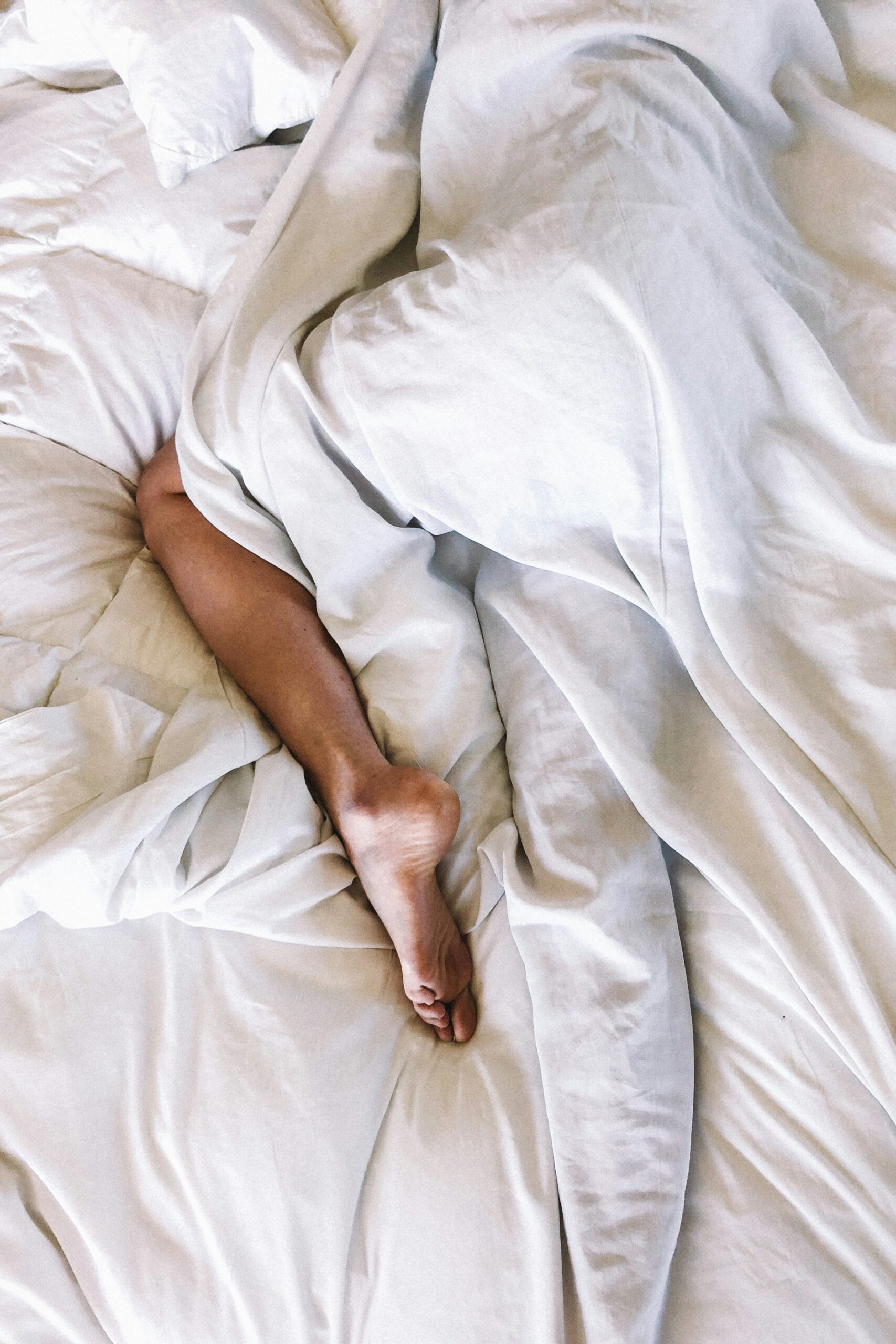 A leg with bare foot seen under crumpled white bedsheets.