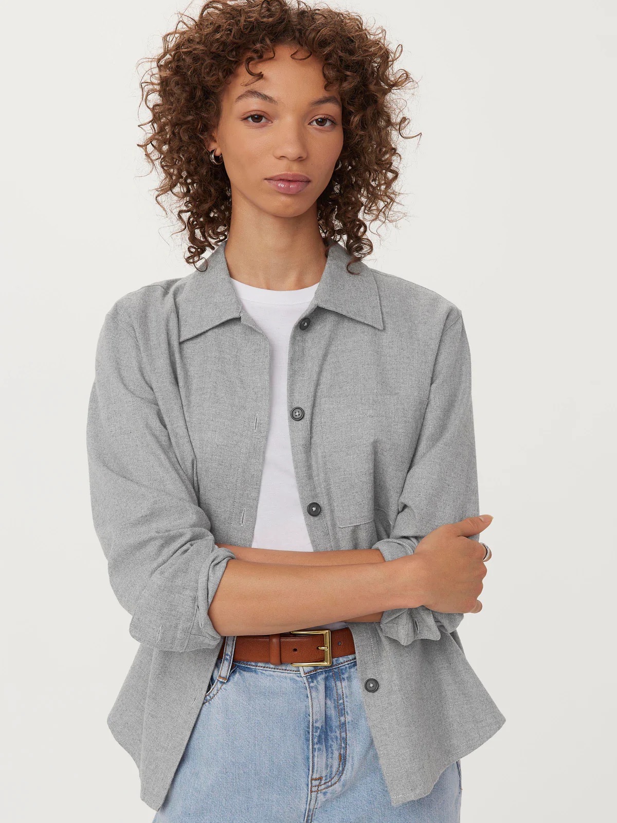 Person with curly hair wears a light gray button-up shirt over a white top and light blue jeans, arms crossed.