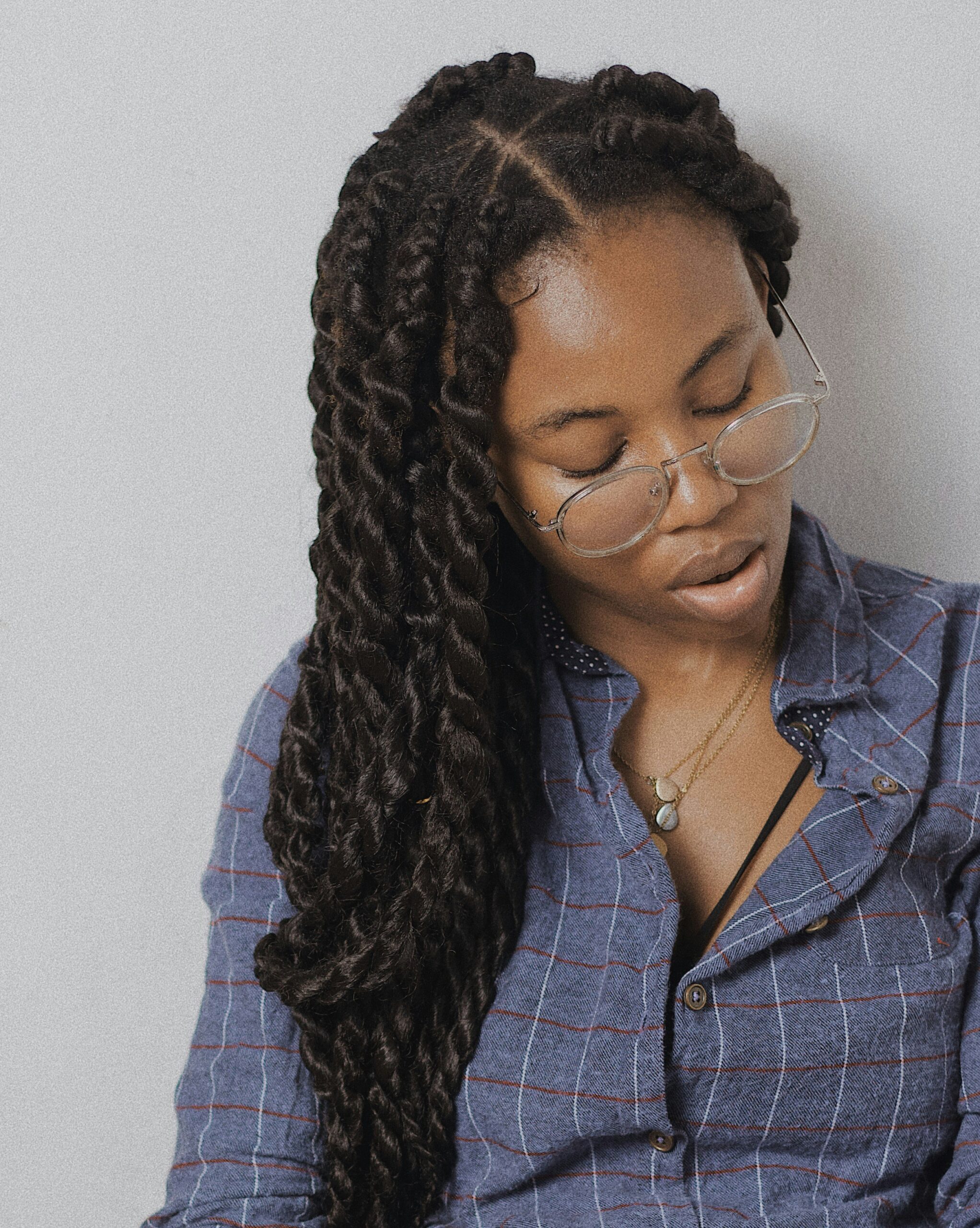 A person with long twisted hair and glasses looks down, wearing a blue checkered shirt and a necklace against a plain background.