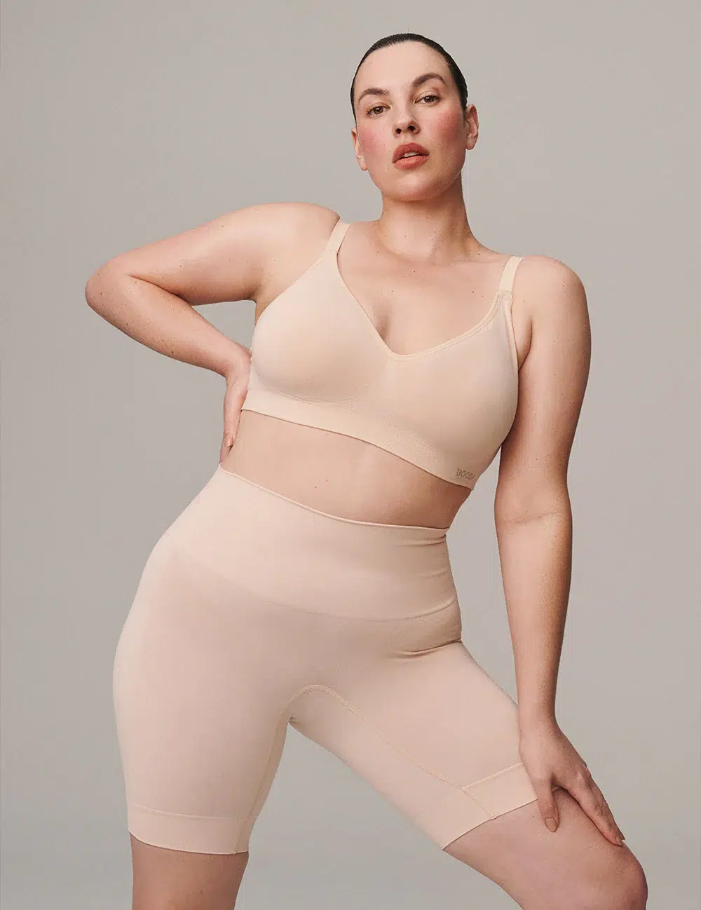 A woman poses in a beige seamless bralette and matching high-waisted biker shorts against a plain background.