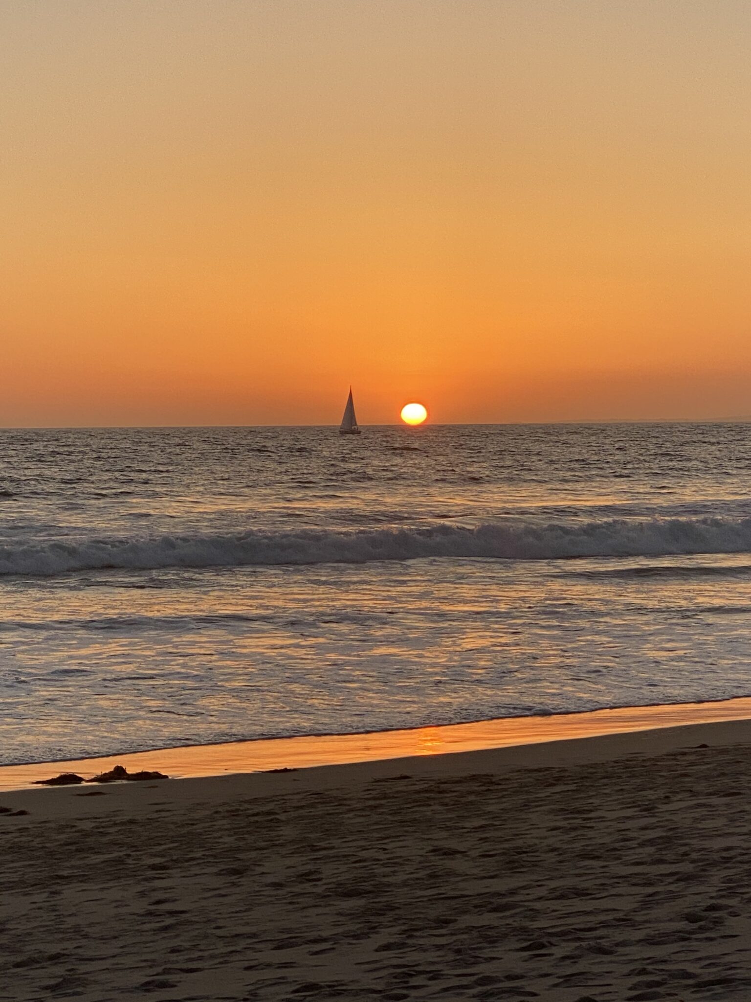 A sailboat passes in front of the setting sun on the ocean, with gentle waves and a sandy beach in the foreground.