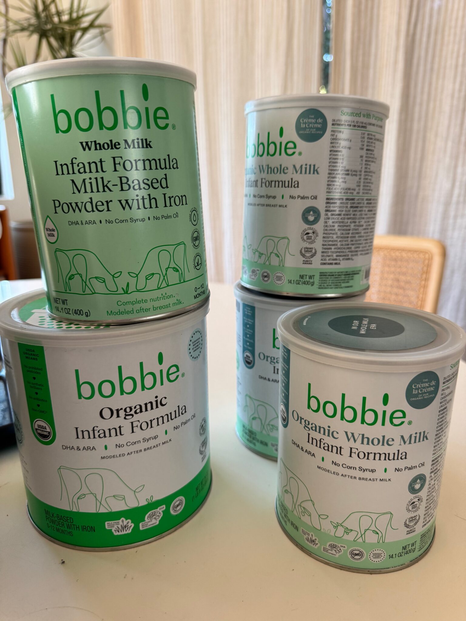 Five cans of Bobbie organic whole milk infant formula are displayed on a white surface in a well-lit room with beige curtains and a wicker chair in the background.