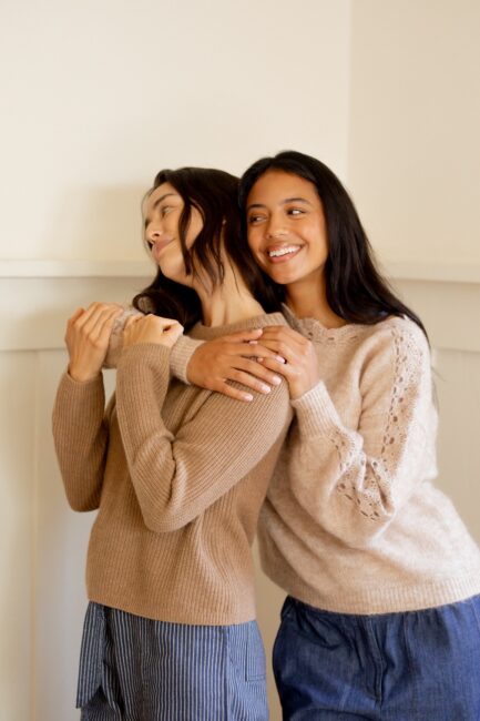 Two women standing close together indoors, both wearing light sweaters and smiling, with one woman embracing the other from behind.