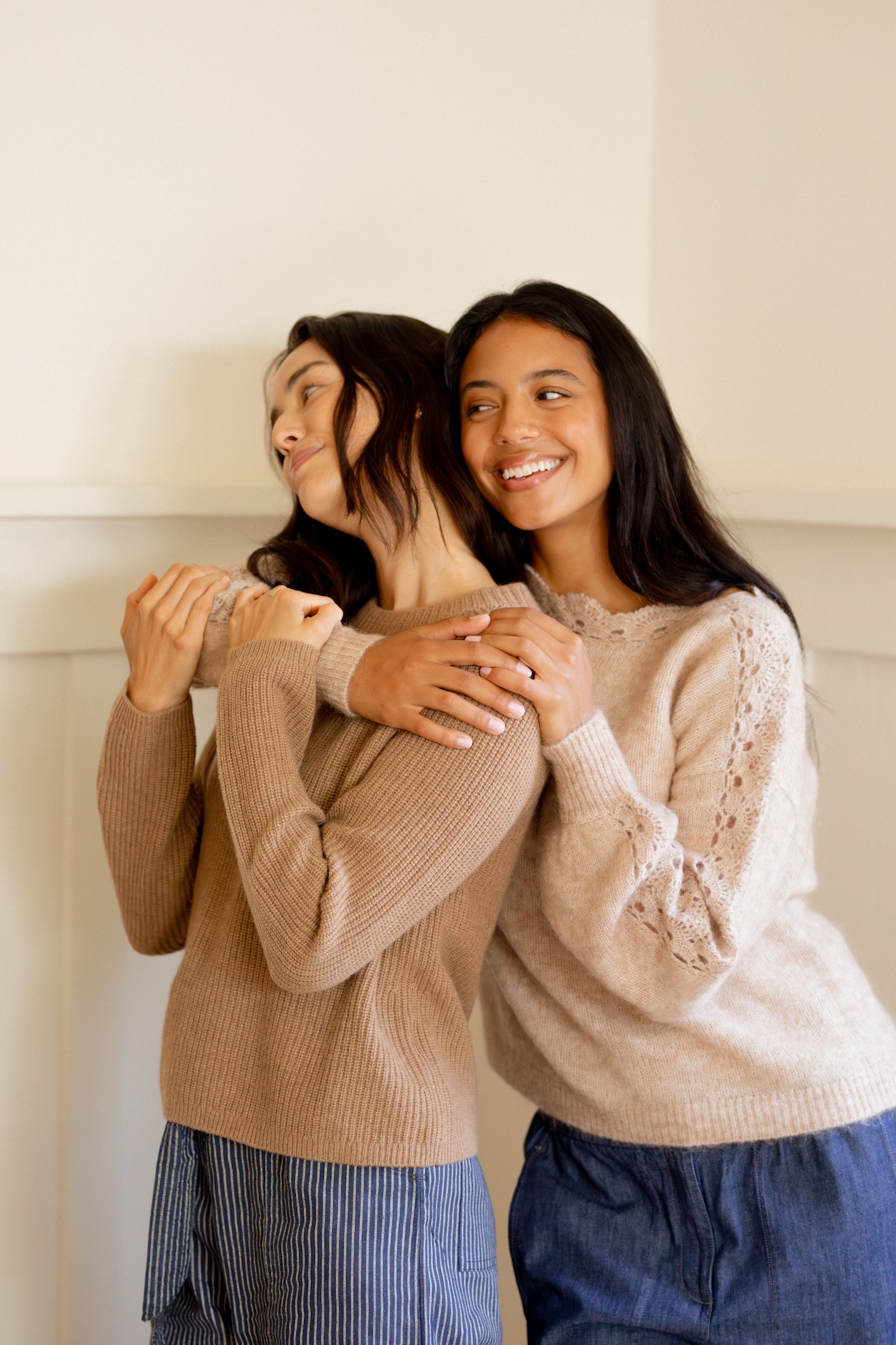 Two women standing close together indoors, both wearing light sweaters and smiling, with one woman embracing the other from behind.