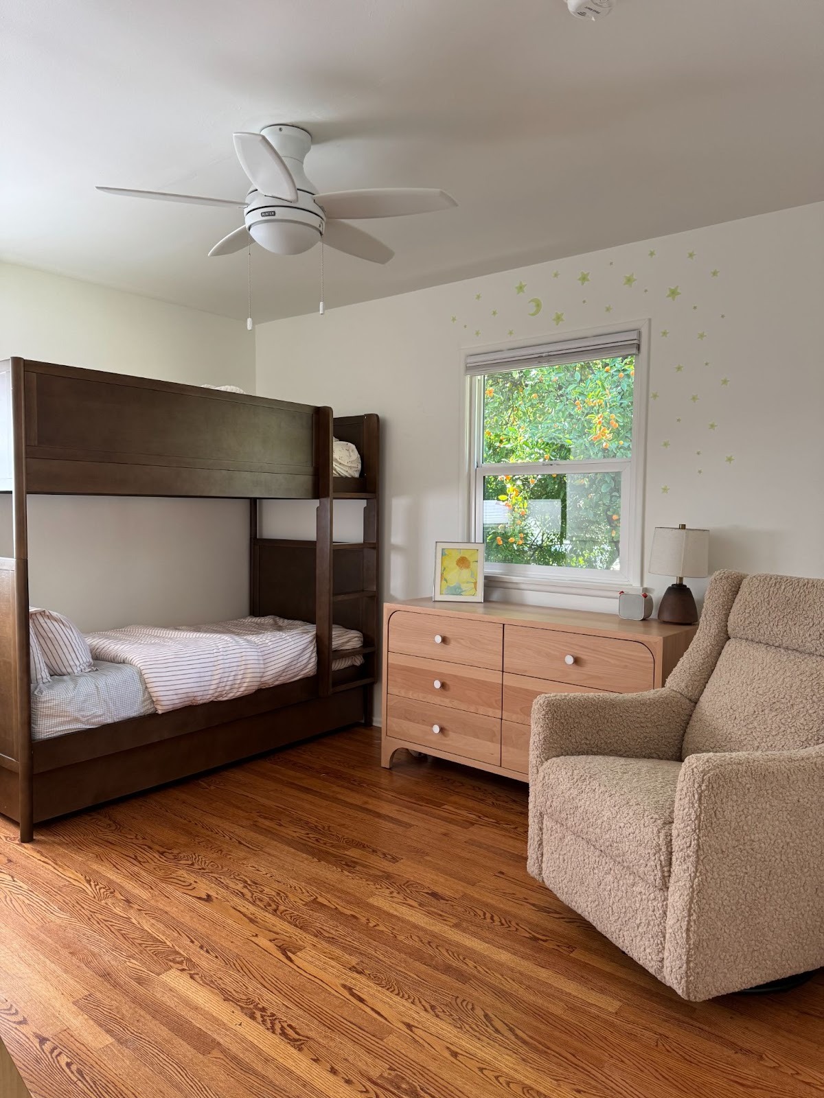 A child's bedroom with a wooden bunk bed, a beige armchair, a light wood dresser, a window with green trees outside, and glow-in-the-dark stars on the wall.