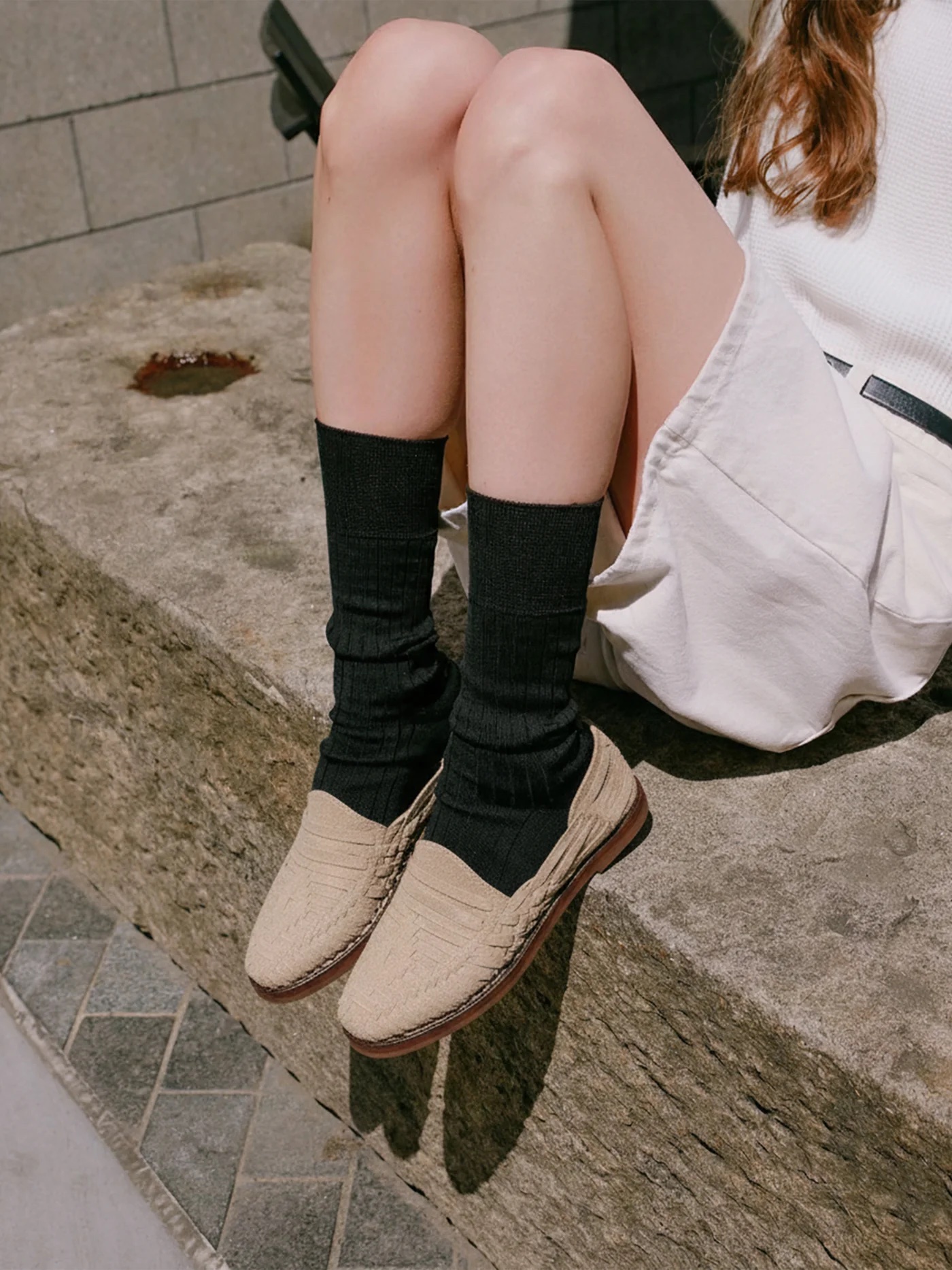 A person sits on a stone ledge, wearing a white skirt, long black socks, and beige huarache sandals. Their hands rest gently on their lap.