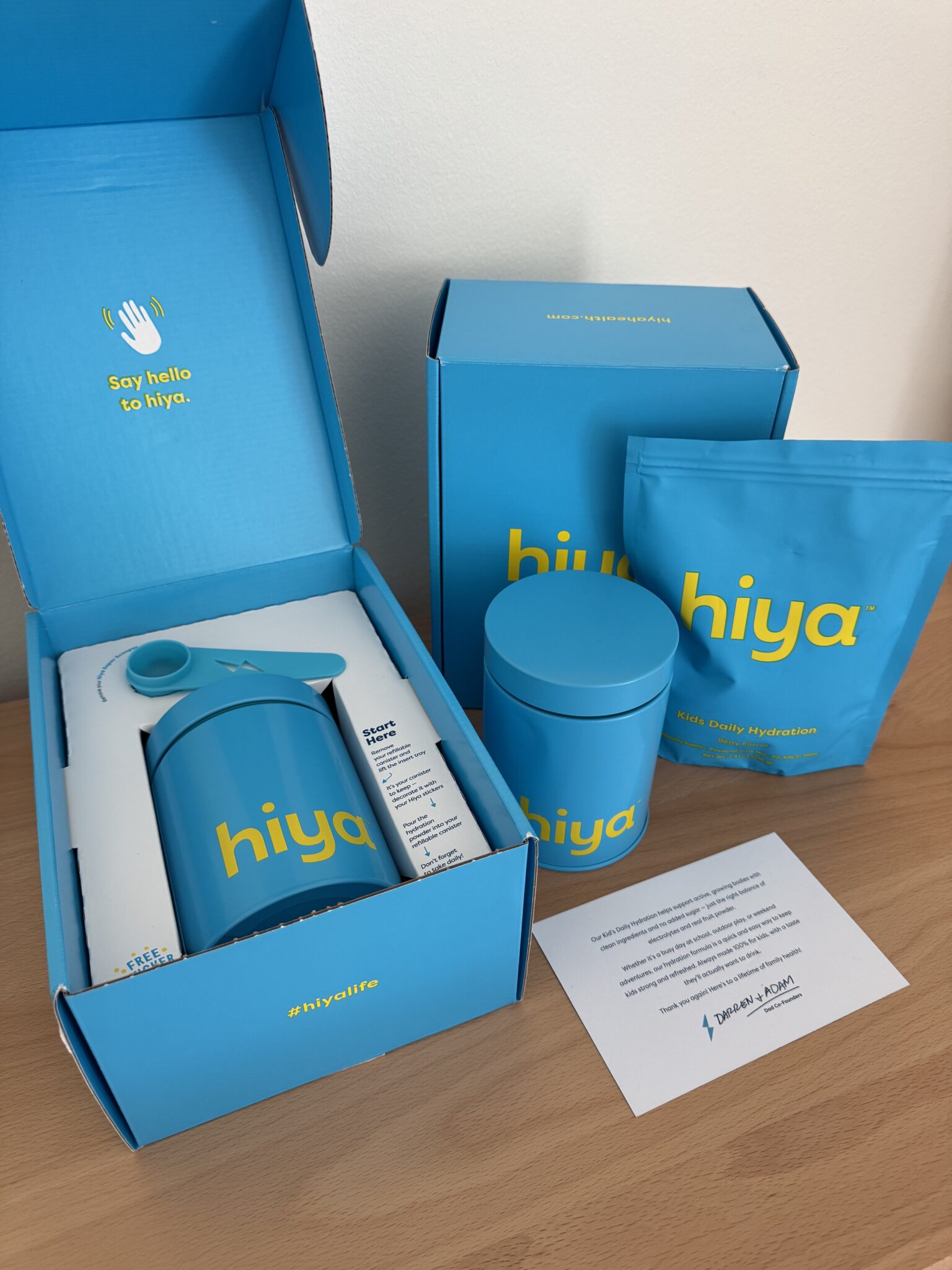 A set of blue hiya-branded hydration products, including a bottle, canister, and pouch, arranged on a wooden surface with blue packaging and an informational card.