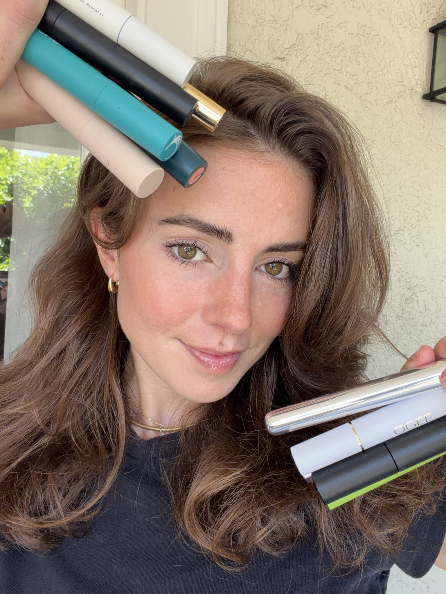 A woman with wavy brown hair holds several mascara tubes near her face and head, smiling slightly at the camera.