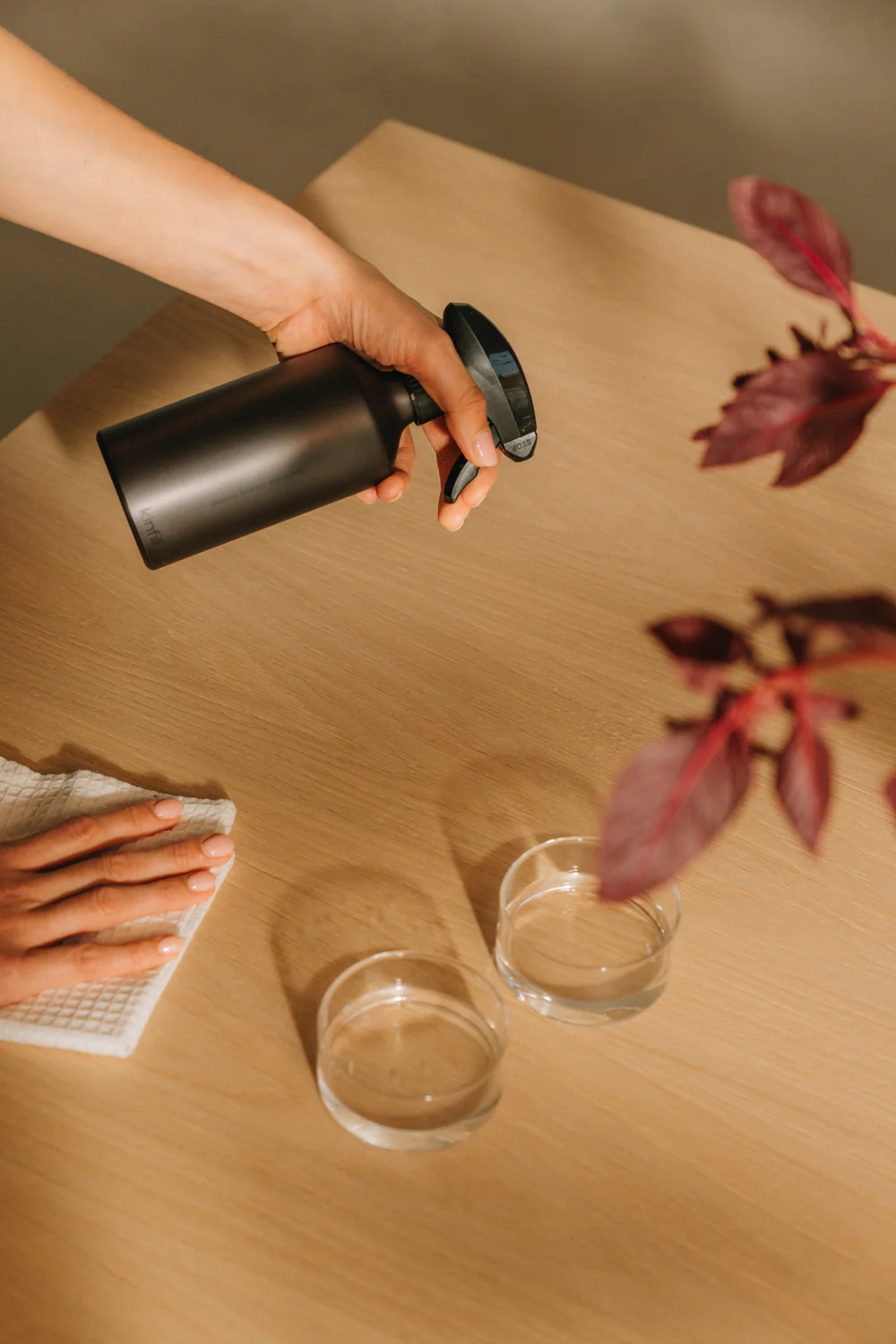 A person sprays a black bottle onto a wooden table while another hand holds a cloth; two glass dishes and a plant are also on the table.