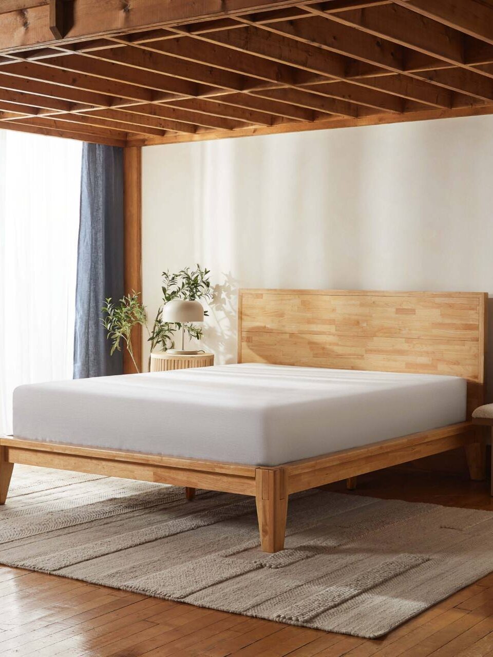 A minimalist bedroom with a wooden bed frame and white mattress, a small plant on a nightstand, and a textured rug on a wooden floor, with sunlight coming through sheer curtains.