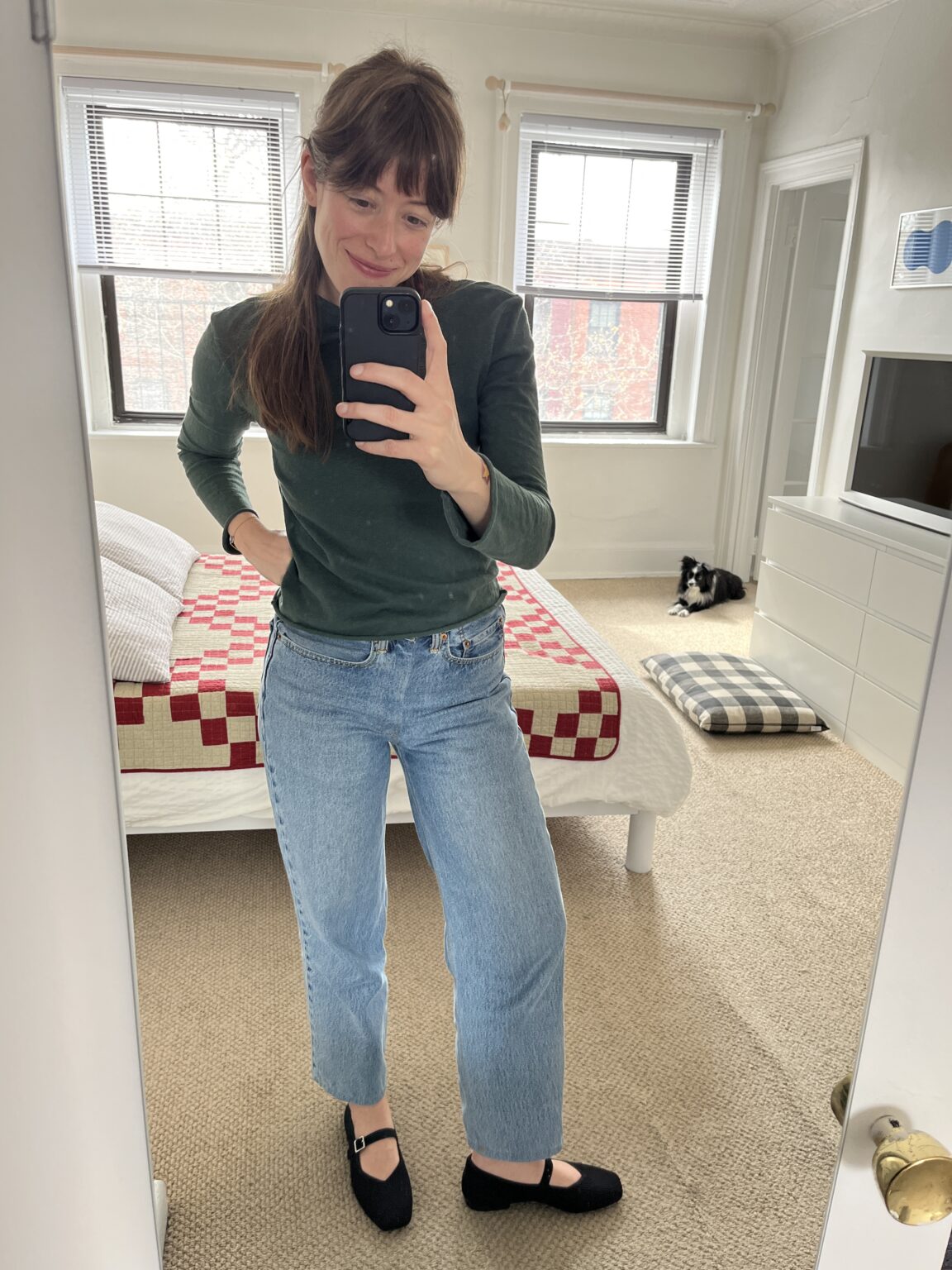 Woman taking a mirror selfie in a bedroom with a made bed, checkered quilt, and a black and white cat sitting near a large plaid pet bed.