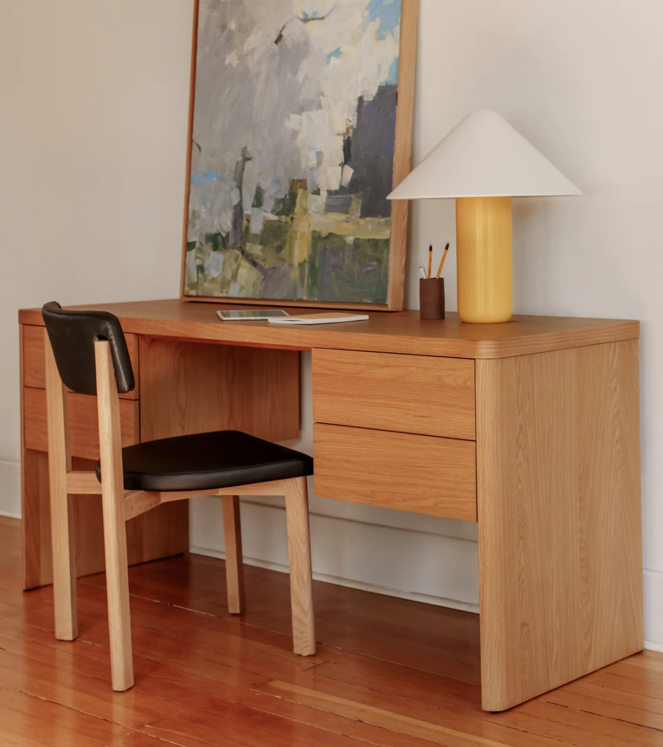 A wooden desk with four drawers holds a lamp, a painting, pencils in a cup, and a closed laptop. A wooden chair with a black seat is placed in front of the desk.
