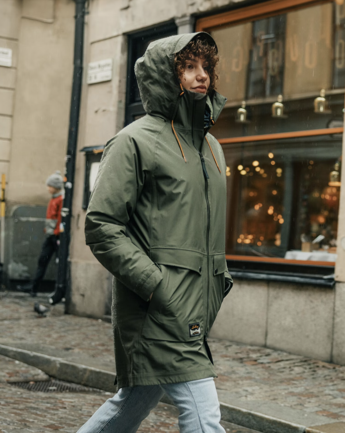 A person wearing a green waterproof raincoat and jeans walks on a cobblestone street in an urban area with a restaurant in the background.