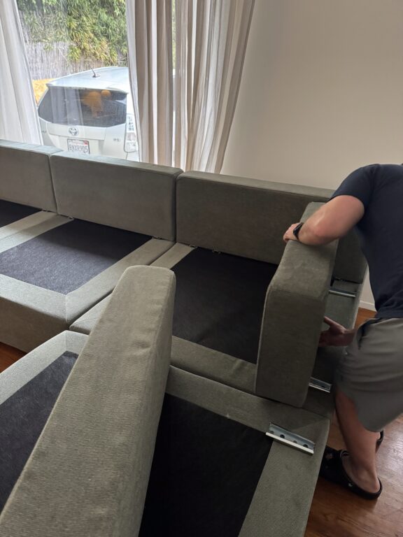 A person in shorts and a t-shirt is assembling or adjusting modular sofa sections in a room with wood floors and large windows.