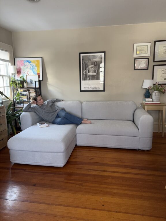 A person lounges on a light gray sectional sofa in a living room with wooden floors, framed art and certificates on beige walls, and books and plants nearby.