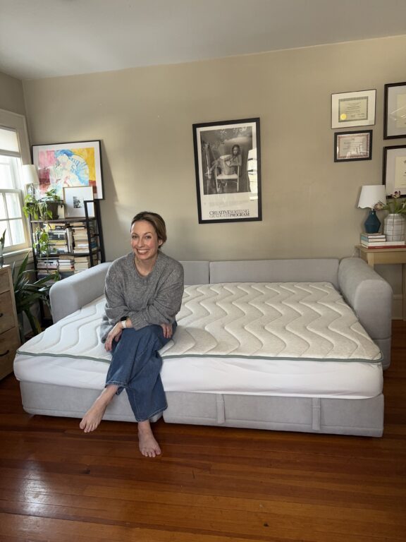 A woman sits barefoot on a gray daybed with a mattress in a cozy, well-lit room decorated with framed art, books, and plants.