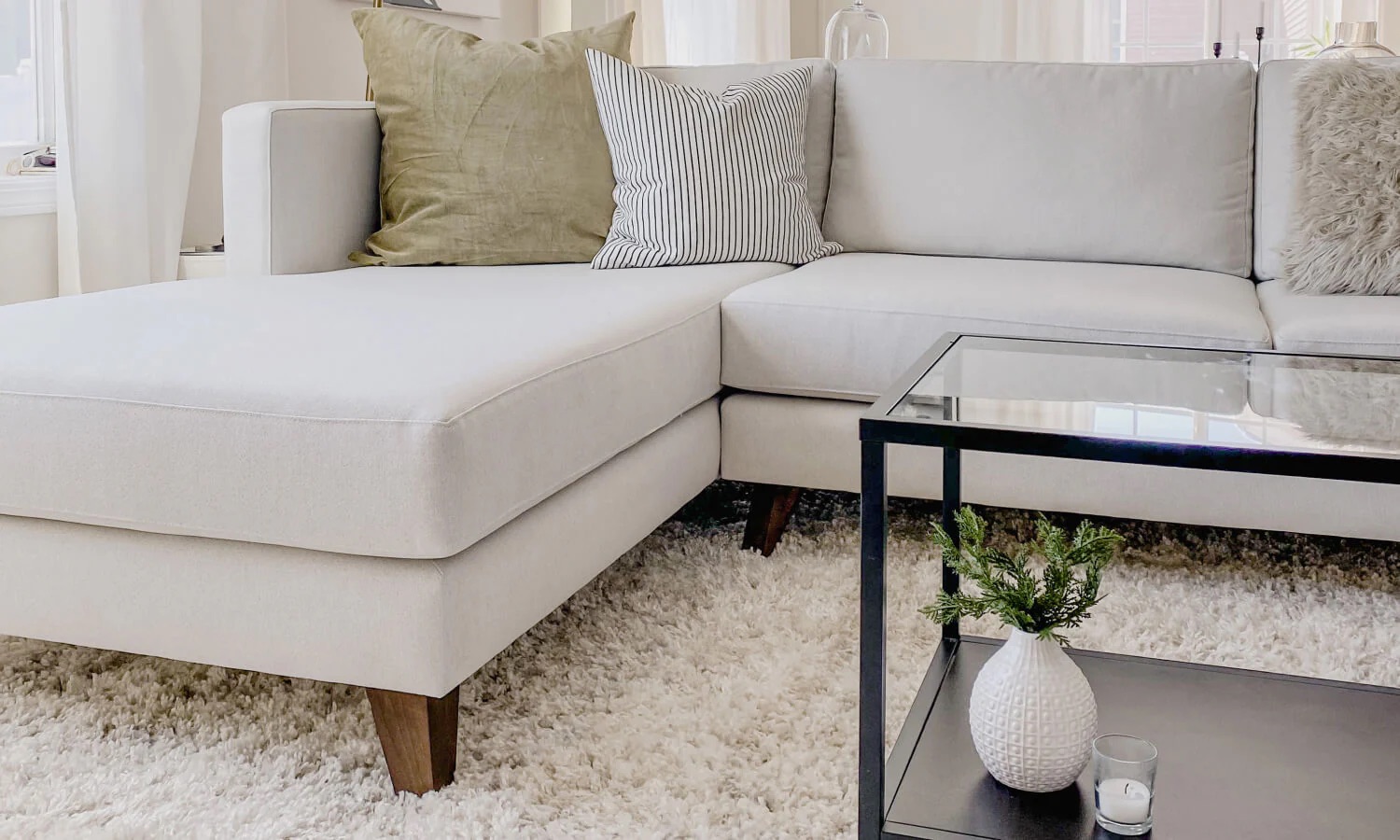 Modern living room with a white sectional sofa, green and striped pillows, glass coffee table, small plant, candle, and a textured white rug.