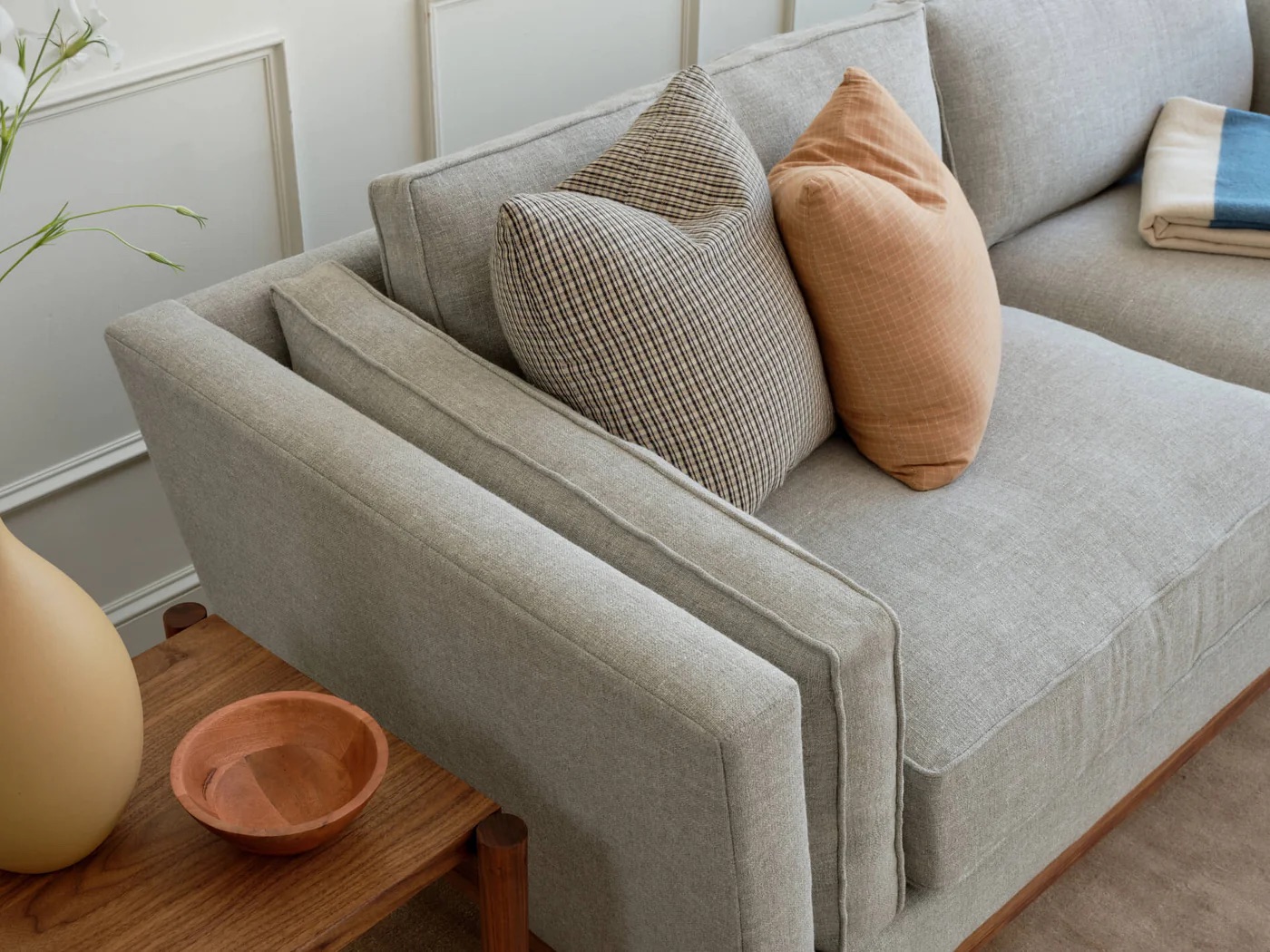 Light gray sofa with two patterned accent pillows in tan and brown, next to a wooden side table with a vase, a small bowl, and a folded blanket on the sofa arm.