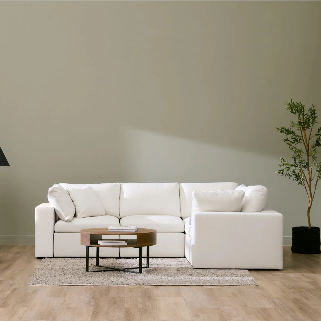 A white sectional sofa sits on a light rug with a round wooden coffee table in front, next to a tall plant, against a pale wall in a minimalist room with wood flooring.