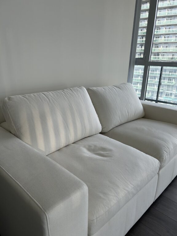 A white two-seater couch sits against a plain wall near a window with blinds, letting in daylight.