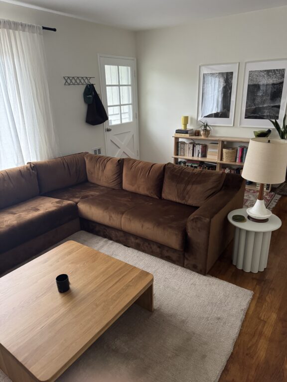 A living room with a brown sectional sofa, light wood coffee table, side table with lamp, bookshelf, and framed art on white walls. White curtains cover a window near the door.