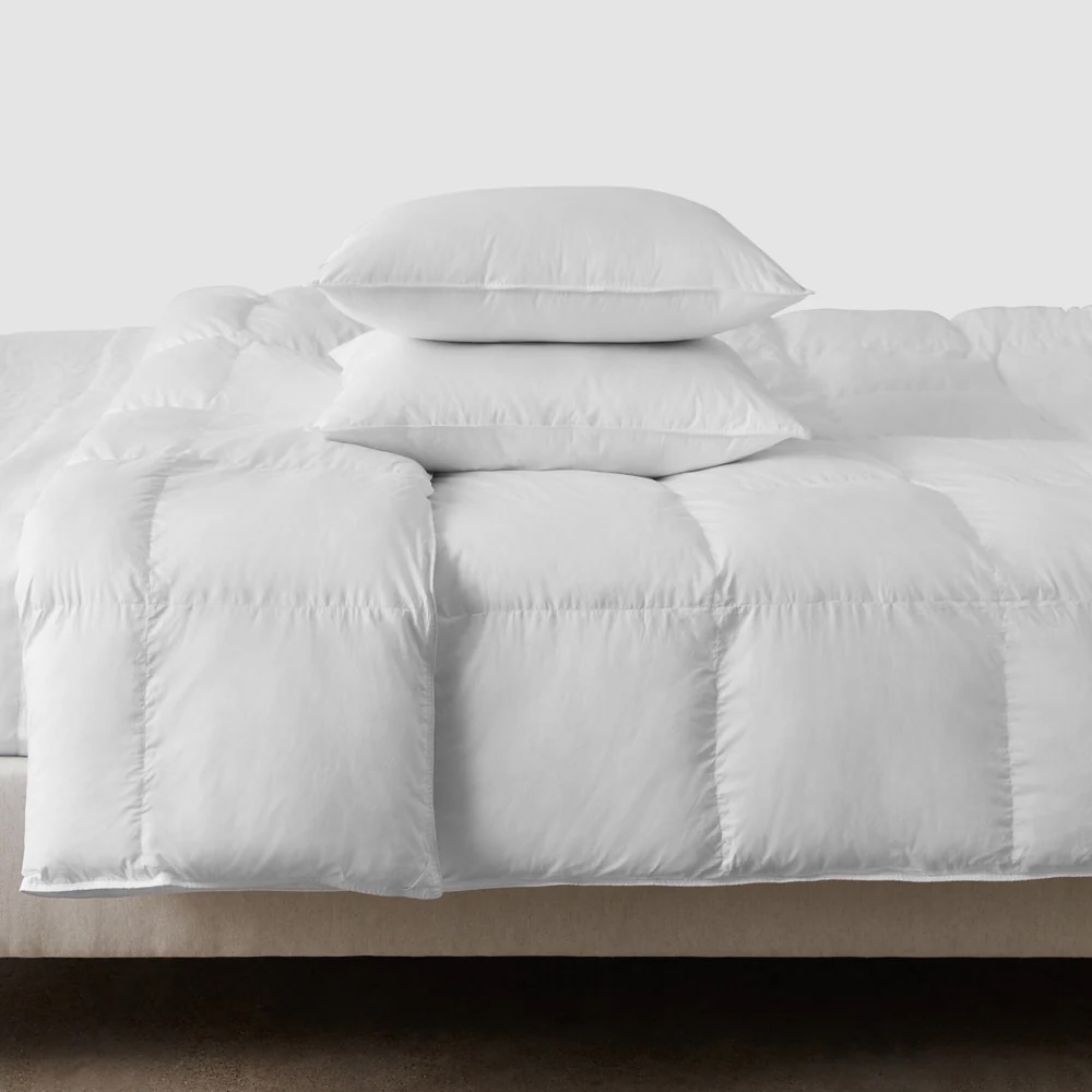 A bed with a white, quilted comforter and two stacked white pillows against a plain background.