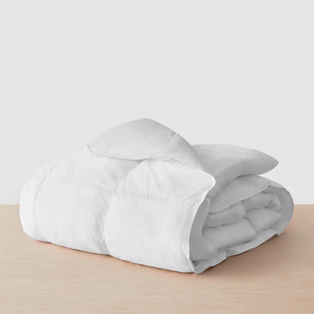 A white, folded comforter is placed on a light wooden surface against a plain, light gray background.