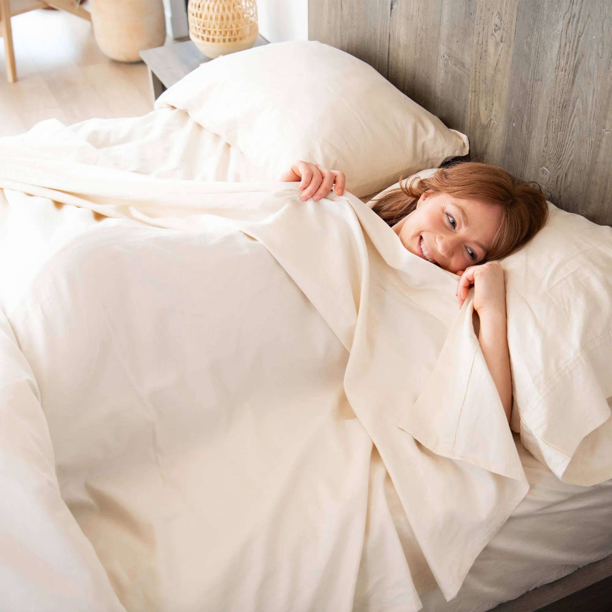 A woman with red hair lies in bed under cream-colored sheets, half-covered, resting her head on a pillow and looking toward the camera.