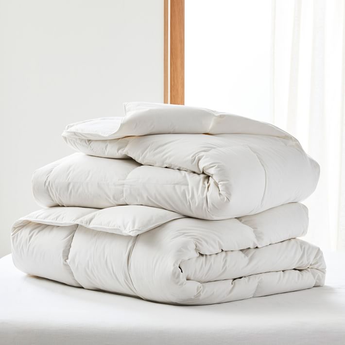 A neatly folded white comforter sits on a bed near a window with natural light and white curtains in the background.