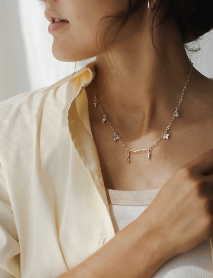 A woman wearing a cream shirt and a white ribbed top displays a silver chain necklace with small charms.