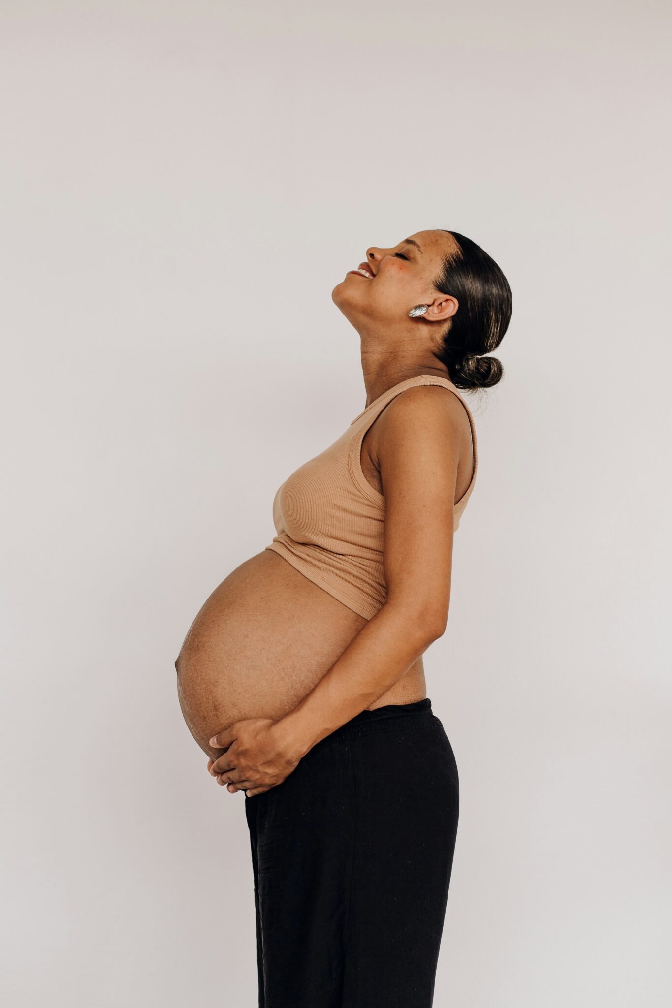 A pregnant woman in a beige sleeveless top and black pants stands in profile, looking upward with a neutral expression.