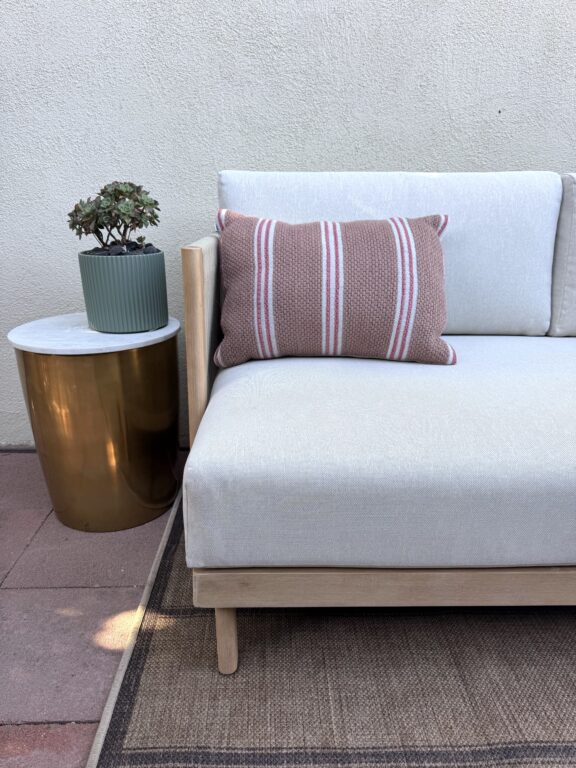 A modern outdoor sofa with a striped pillow sits next to a round gold side table holding a potted succulent, against a light-colored wall.