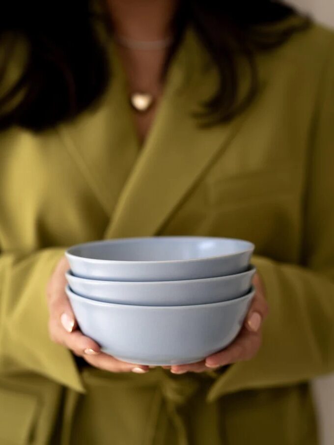 Person in a green blazer holding a stack of three light blue bowls with both hands.