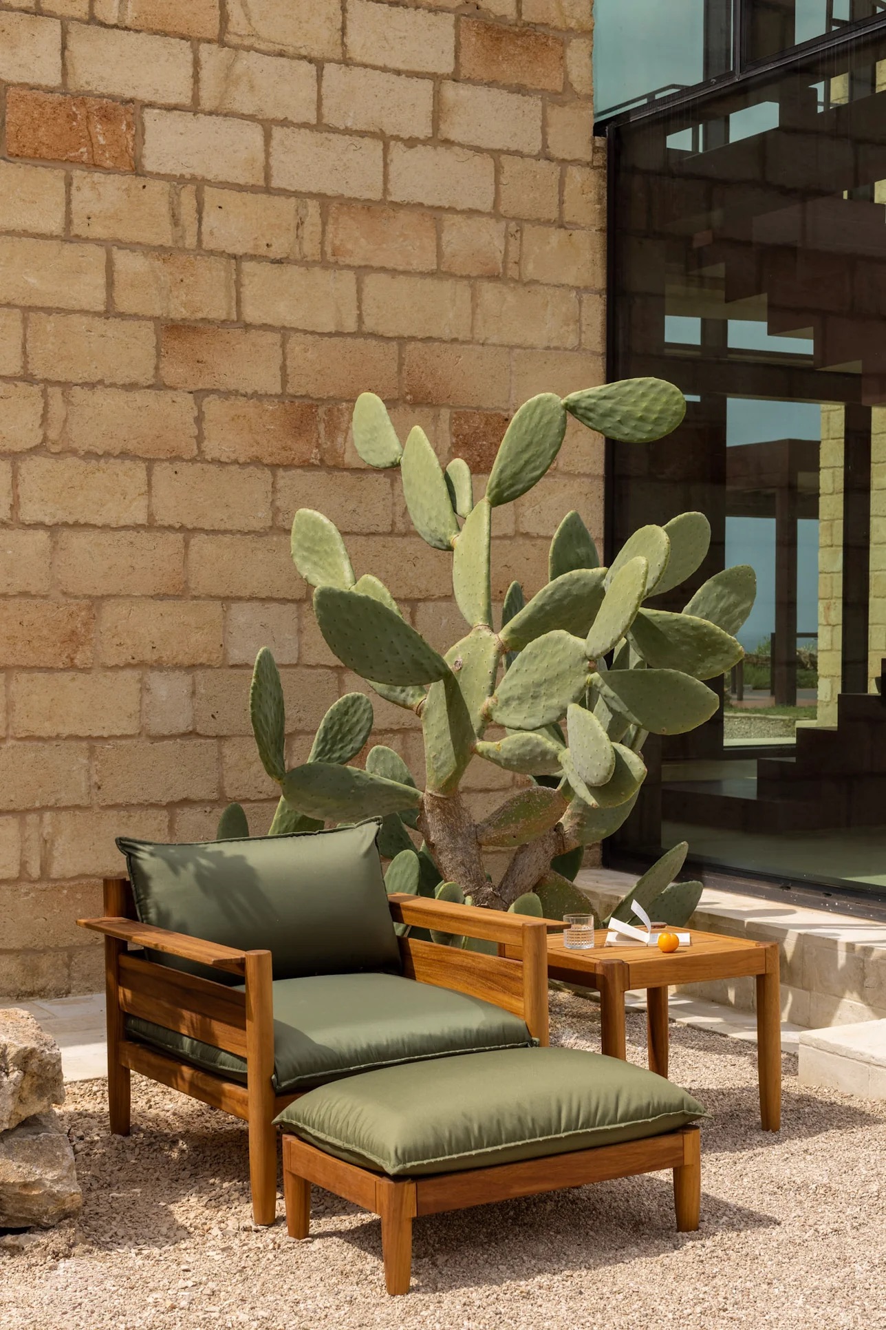 A wooden lounge chair with green cushions and a matching table sit on gravel beside a stone wall, near a large prickly pear cactus.