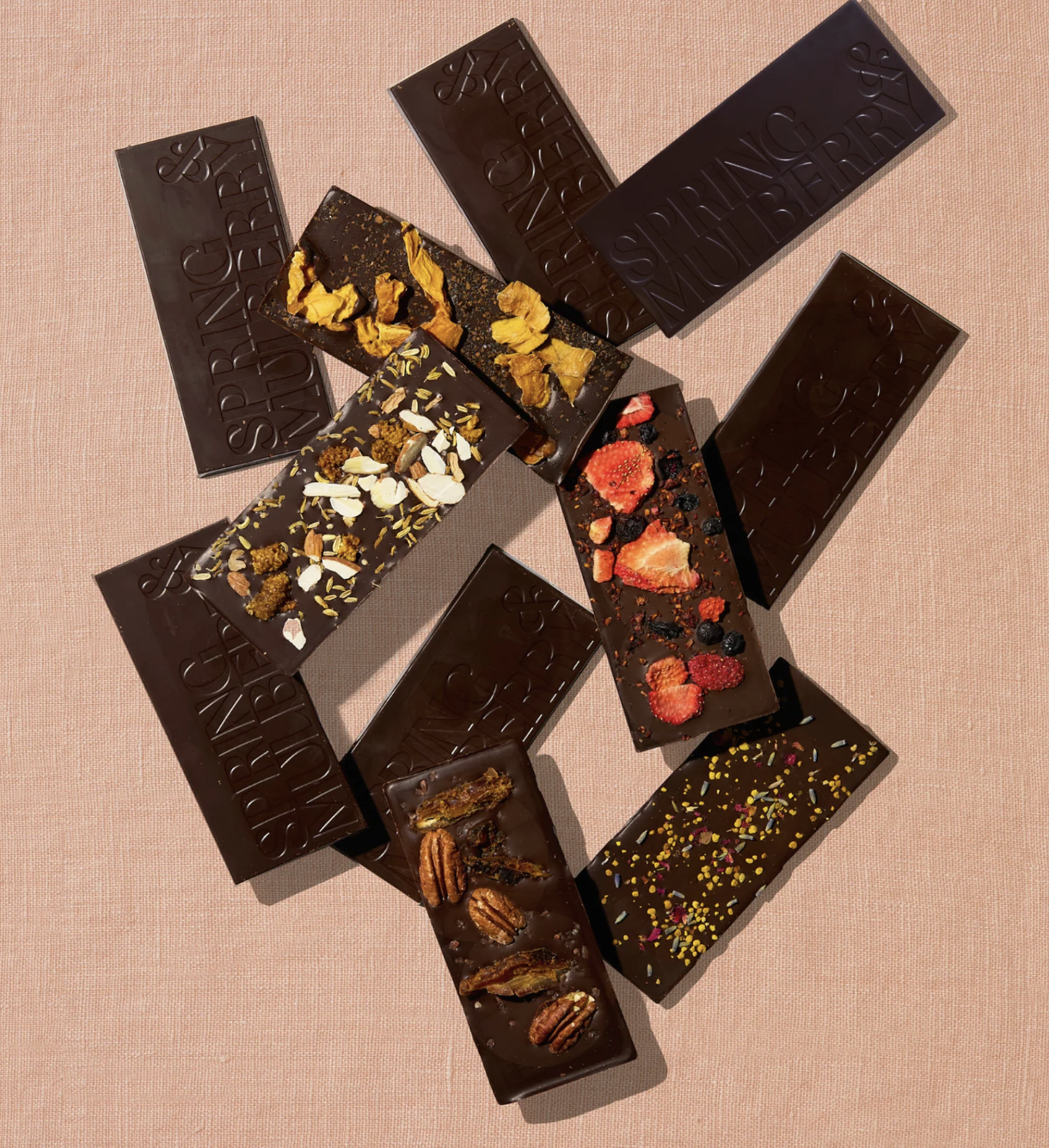 Assorted dark chocolate bars, some topped with nuts, dried fruit, cornflakes, or sprinkles, displayed on a pink textured surface.