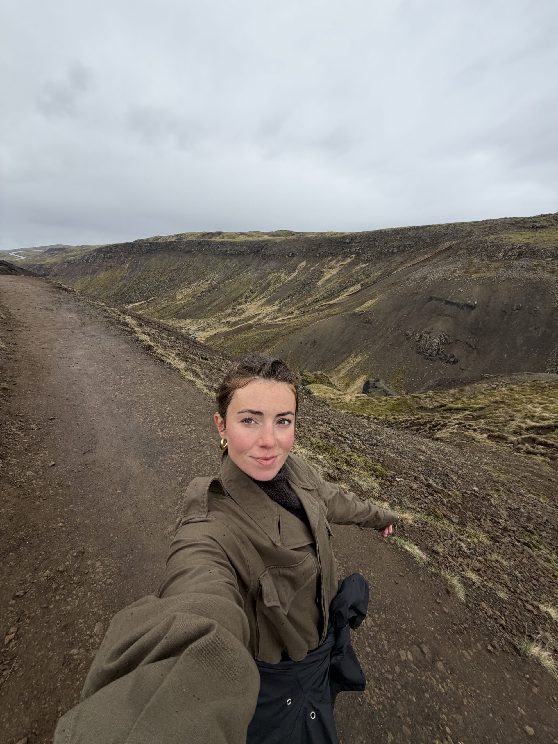 Person taking a selfie on a dirt path in a hilly, rocky landscape under an overcast sky.
