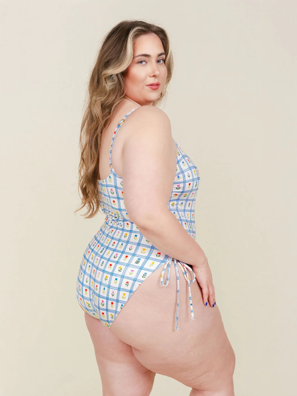A woman with long wavy hair poses in a light blue patterned one-piece swimsuit with side ties, standing against a plain beige background.
