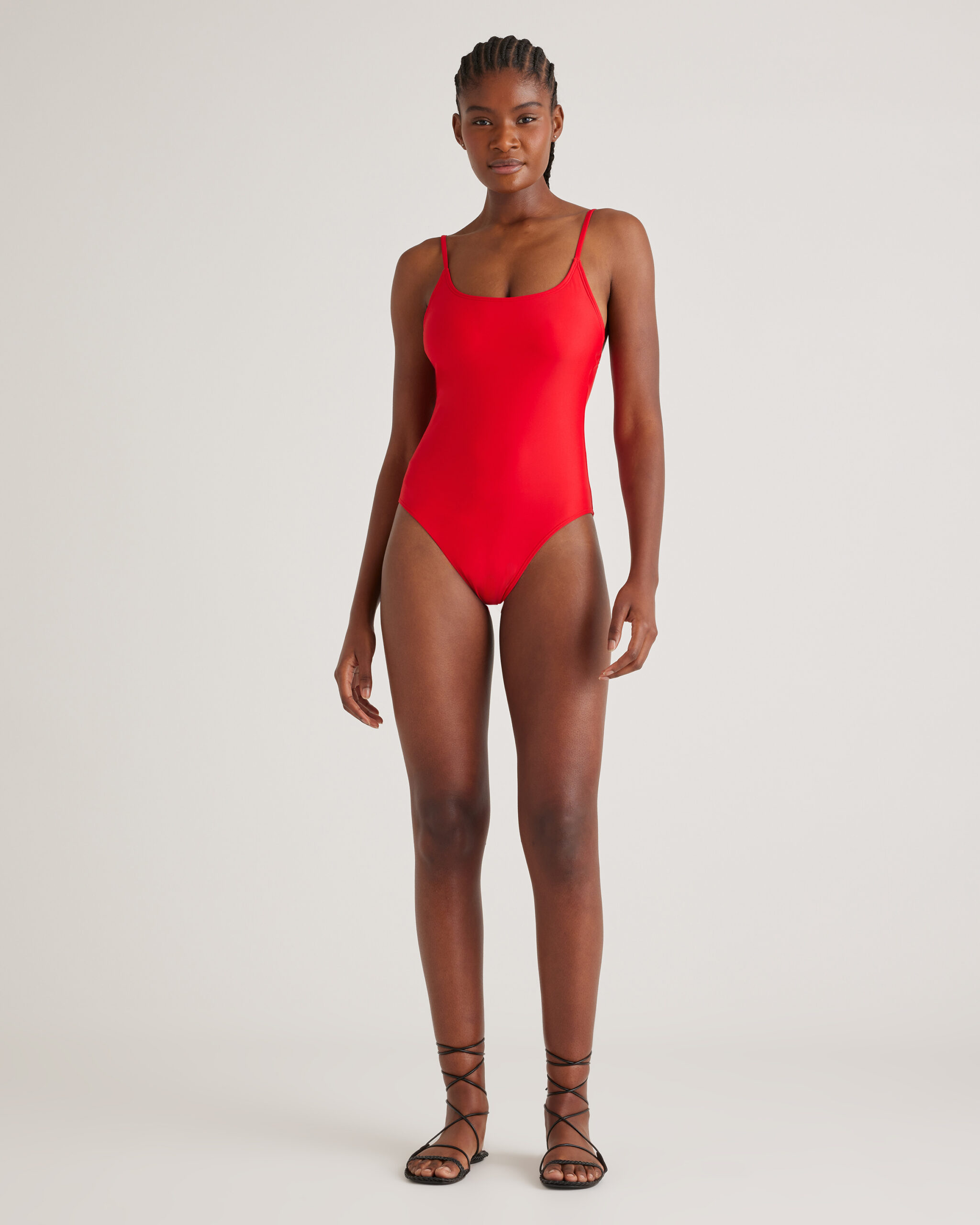 A person stands facing forward, wearing a red one-piece swimsuit and black strappy sandals against a plain light background.