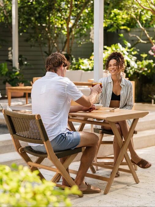 Two people sit at a wooden table outdoors near a pool, smiling and holding hands, surrounded by green plants and patio furniture.