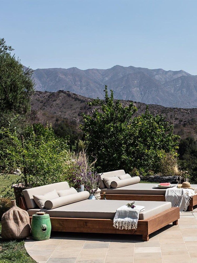 Two outdoor lounge beds with cushions and towels are positioned beside a pool, with mountains and greenery visible in the background under a clear sky.