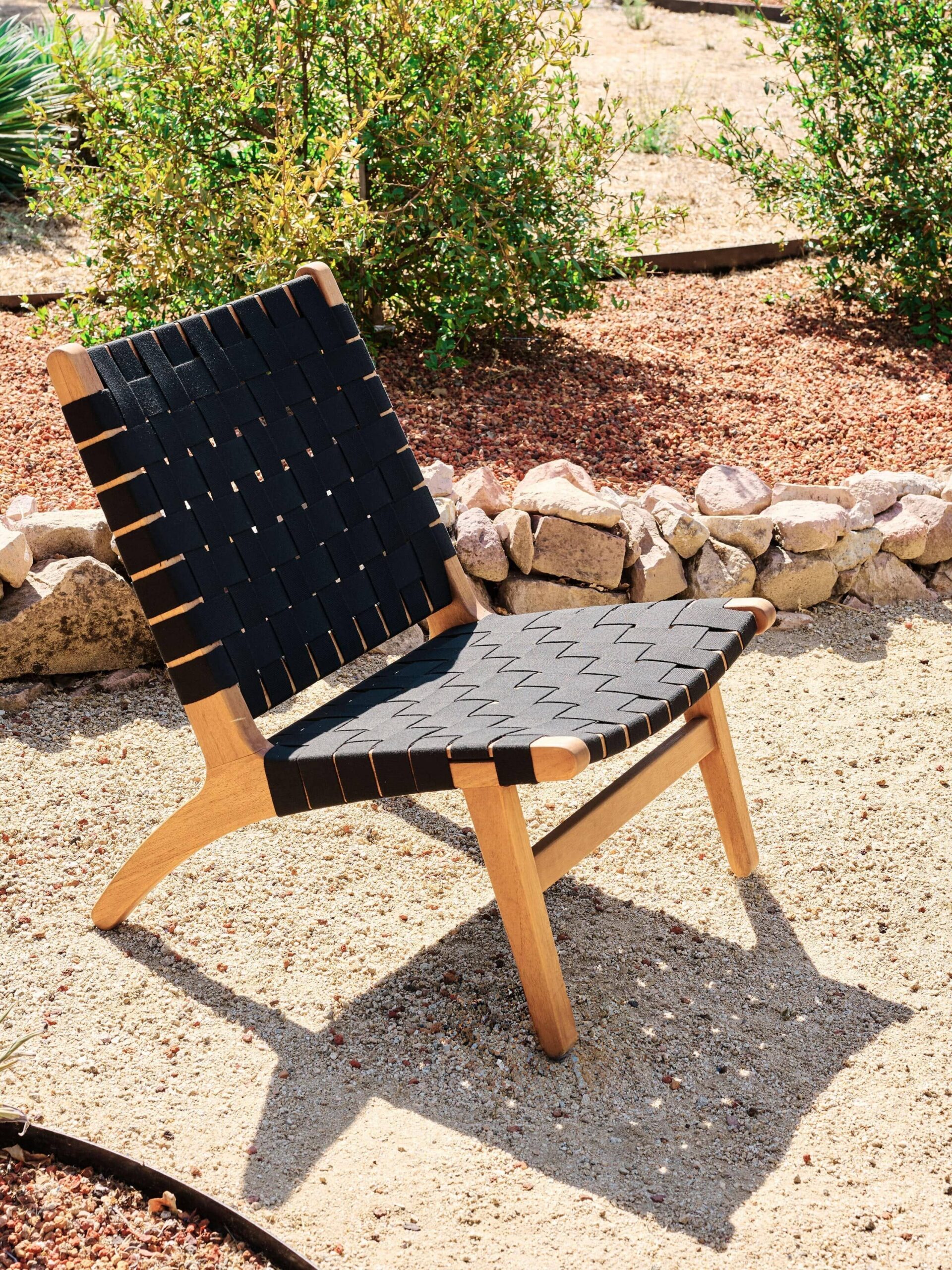 A wooden chair with a woven black seat and backrest is placed outdoors on a sandy surface, surrounded by rocks and plants.