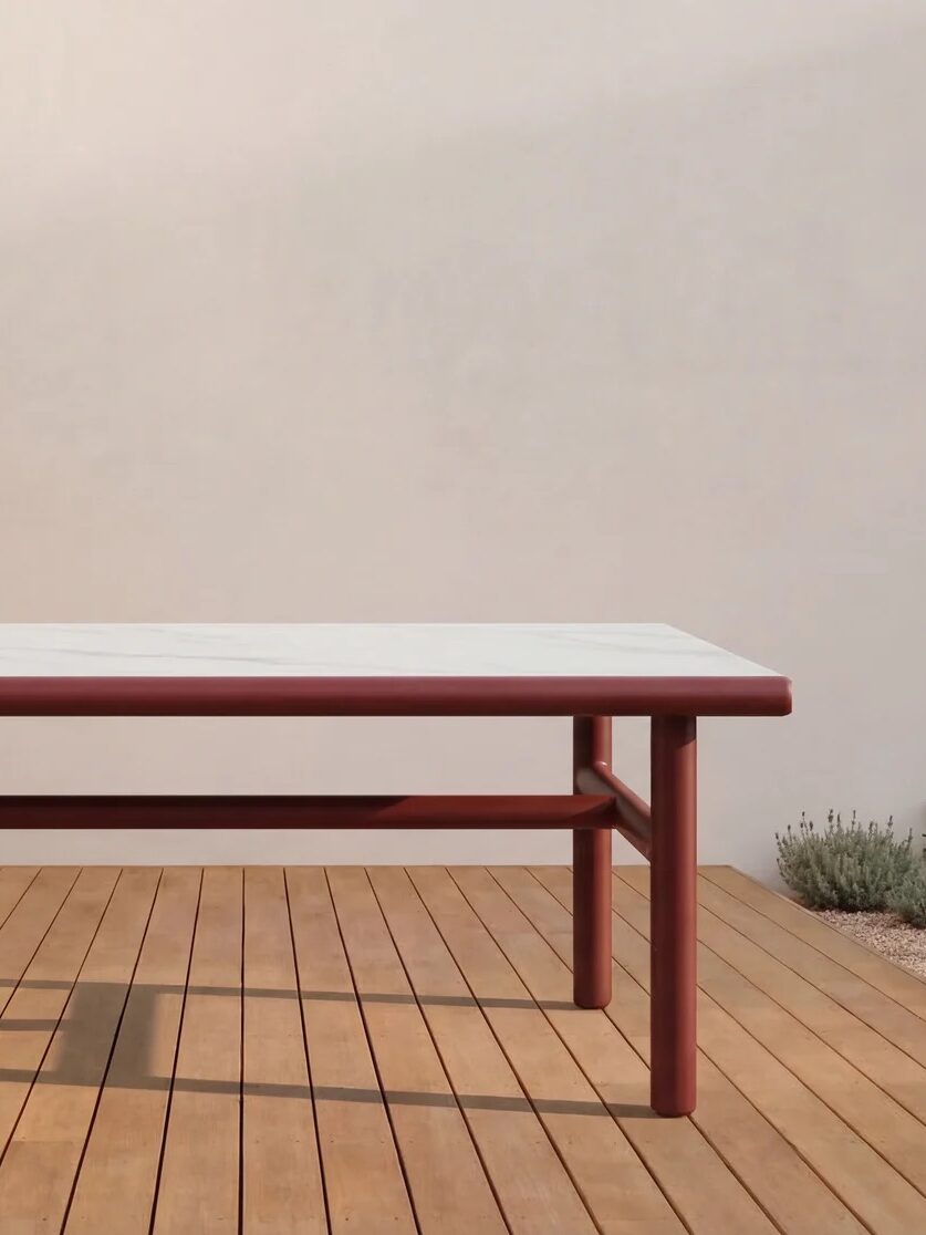 A rectangular table with a white top and red legs stands on a wooden deck, surrounded by small plants and next to a light-colored wall.